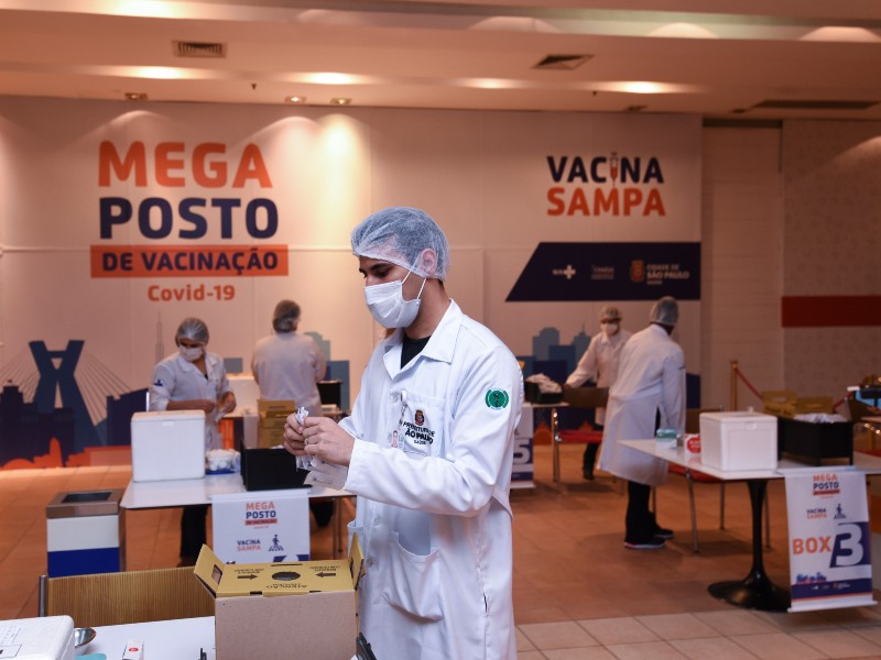 Foto de um dos megapostos de vacinação. À frente, um profissional da saúde manipula um material de vacinação. Na frente dele há uma mesa com uma caixa em cima. Ao fundo aparecem outros quatro profissionais da saúde trabalhando, outras mesas com insumos para vacinação e também uma parede com o letreiro megaposto de vacinação Covid-19 Vacina Sampa